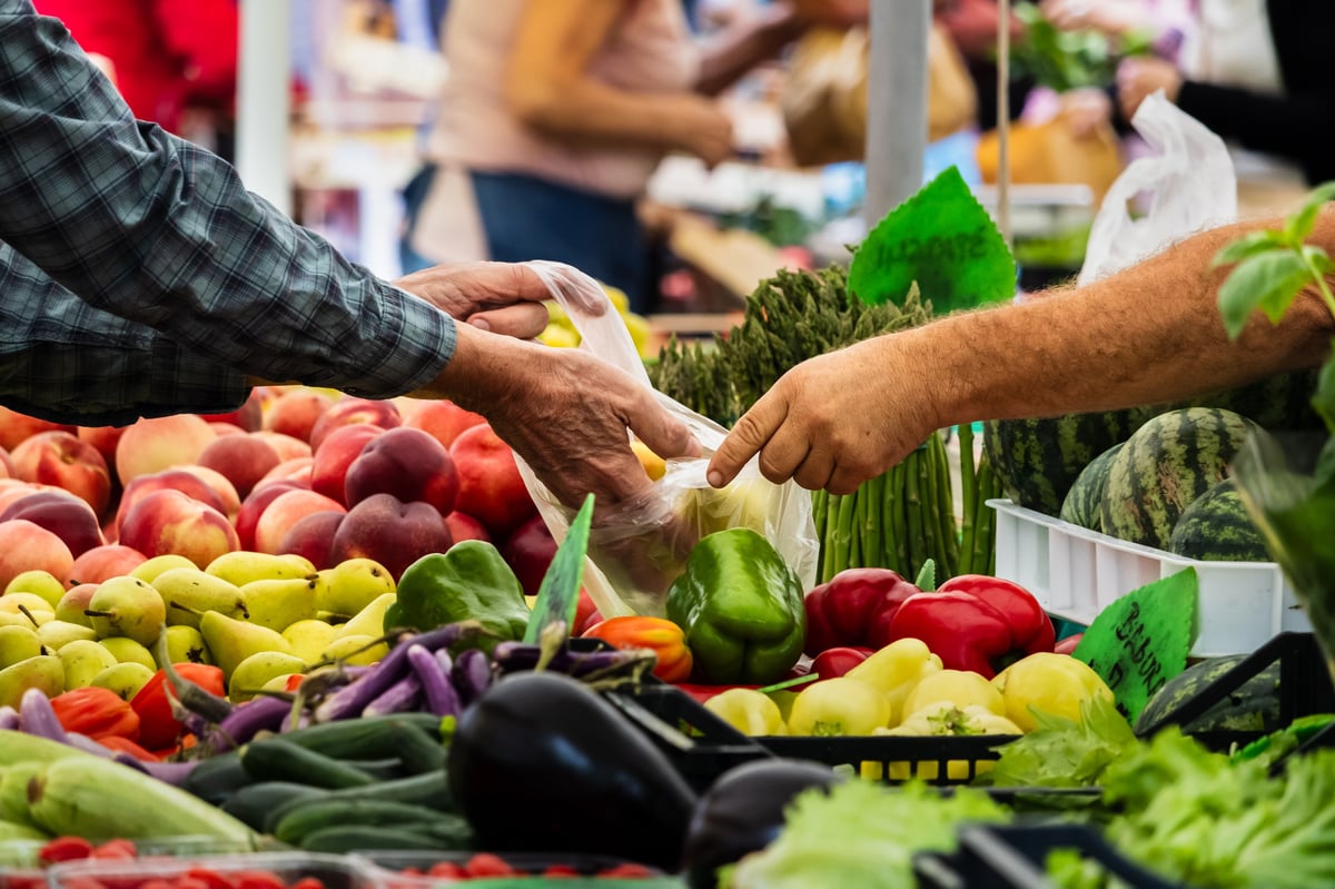 A buyer and a vendor exchanging fresh vegetables at a busy farmers market, with peppers, peaches, eggplants and other seasonal produce in the foreground