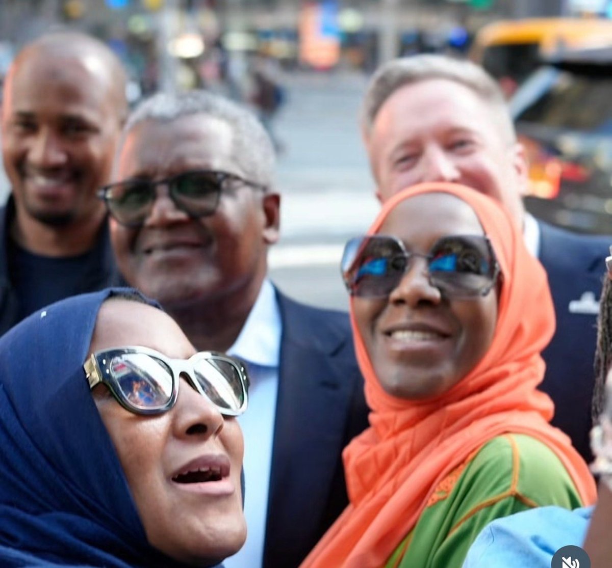 Group of five people wearing sunglasses smiling at a street event with yellow taxi and buildings in the background