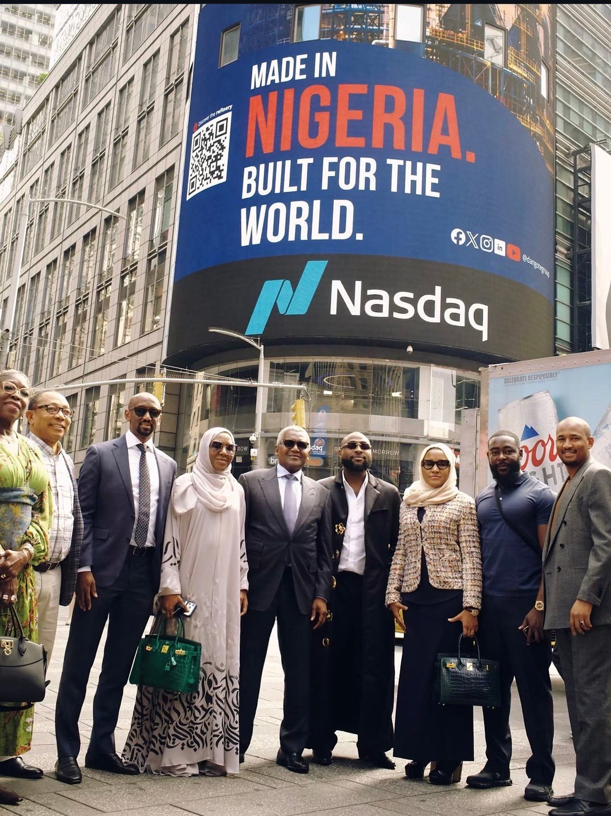 A group of formally dressed people stand in front of a large blue Nasdaq billboard reading Made in Nigeria. Built for the World. in an urban setting.